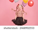 A happy cute little girl in shiny party clothes and with long pigtails tied to colorful balloons smiles cheerfully. Portrait on a pink studio background. Festive kids mood. 