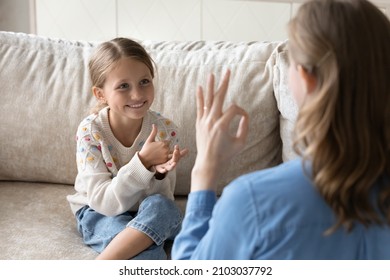 Happy cute little kid girl with hearing disability using sign language, communicating with caring mother or professional female speech therapist, sitting together on comfortable sofa at home. - Powered by Shutterstock