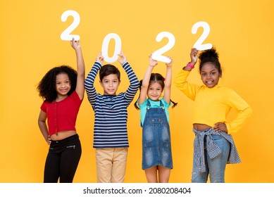 Happy Cute Little Kids Smiling And Holding 2022 Numbers Isolated On Yellow Studio Background For New Year Concepts