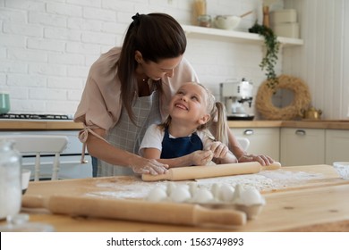 Happy Cute Little Preschool Daughter Enjoying Helping Smiling Mother Preparing Homemade Pastry At Kitchen. Smiling Small Kid Girl Likes Rolling Out Dough, Looking At Joyful Proud Young Mom In Apron.