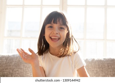 Happy Cute Little Vlogger Waving Hand Saying Hello Hi Looking At Camera Talking To Webcam, Smiling Kid Child Girl Making Online Video Call Recording Vlog Sitting On Sofa At Home, Portrait