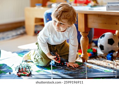 Niño preescolar lindo feliz que juega con los coches de carreras en la pista de carreras, en interiores, con los coches. Niño emocionado que se divierte con el juego interior de la carrera
