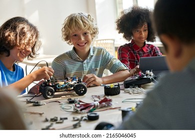 Happy Cute Schoolboy Looks At Camera Sits At Desk With Group Of Diverse Middle School Students Building Innovative Robot Making Software Engineering At Science Lab Class. STEM Technologies Concept.