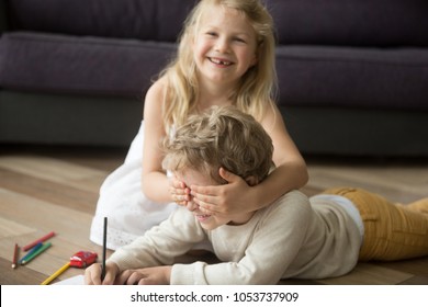 Happy cute siblings enjoying playing peek a boo, kid girl sister laughing closing brothers eyes with hands while child boy drawing on warm wooden floor, children having fun at home together concept
