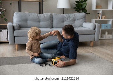 Happy cute small kid son bumping fists with caring millennial father, finishing repairing favorite toy car with screwdriver, having fun improving skills sitting on floor carpet together at home. - Shutterstock ID 2111421614
