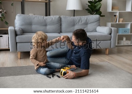 Happy cute small kid son bumping fists with caring millennial father, finishing repairing favorite toy car with screwdriver, having fun improving skills sitting on floor carpet together at home.