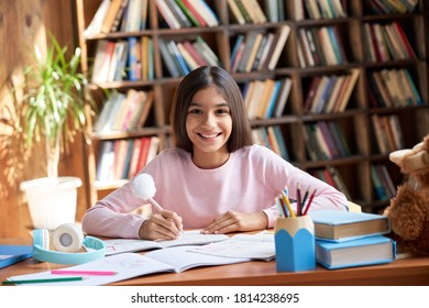 Happy Cute Smart Hispanic Indian Preteen Kid Girl Student, Latin Child Primary School Pupil Studying At Table At Home, Learning Sitting At Classroom Desk Looking At Camera, Schoolgirl Portrait.