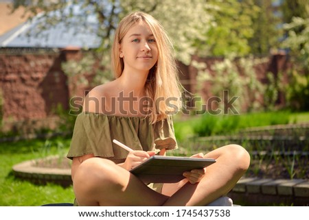 Happy and cute teen girl draws sketches on a tablet sitting in the yard, in the garden on the lawn in Sunny weather.