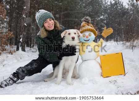 Happy cute teenager girl, white dog, cheerful snowman in yellow knitted hat, jacket, mittens, medical mask with smile painted on it in a snowy forest. life during COVID 19 pandemic. Winter activities