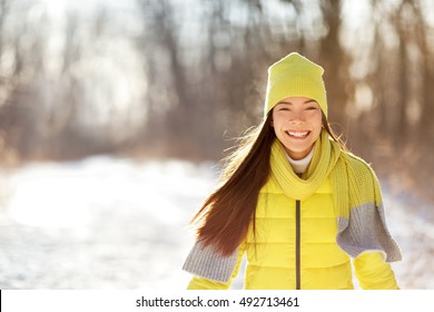 Happy Cute Winter Girl Smiling In Snow Forest. Portrait Of Asian Woman Happy Playful Outdoors With Healthy Smile On Sunny Wintertime Day Wearing Yellow Outerwear Beanie Knit Hat And Scarf.