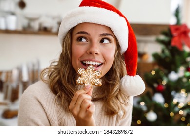 Happy cute woman in santa claus hat smiling and eating Christmas cookie in cozy kitchen