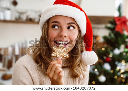 Happy cute woman in santa claus hat smiling and eating Christmas cookie in cozy kitchen