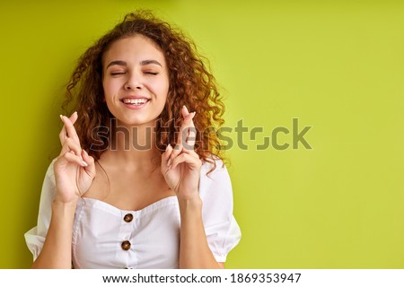 happy cute young woman with curly hair keeps fingers crossed, luck concept. female in blouse is wishing for good luck, isolated over green background