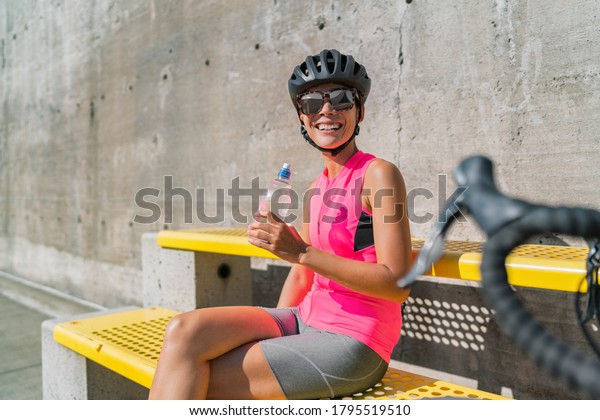 Happy cyclist drinking water\
bottle, tired from heat exhaustion, taking a break during biking\
ride in city. Woman resting by road bike hydrating on summer\
travel.