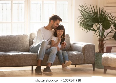 Happy Dad Embracing Little Kid Daughter Enjoy Using Smartphone Gadget Sitting On Couch At Home, Cute Child Girl Holding Phone Device Showing Father New Mobile Apps On Cellphone Having Fun Together