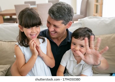 Happy Dad Enjoying Time With Kids, Waving Hand At Camera. Joyful Father And Two Children Sitting On Couch In Living Room Together. Leisure Time With Family Concept