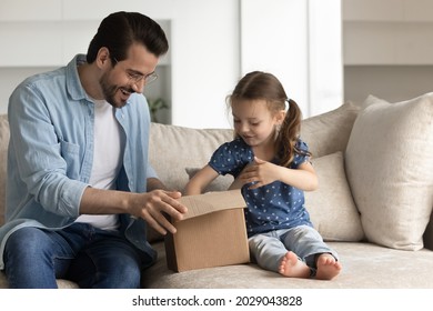 Happy Dad And Excited Daughter Girl Unboxing Parcel With Surprising Gift, Opening Cardboard Box, Looking Inside Container, Producing Goods. Father And Kid Receiving Purchase Via Delivery Service