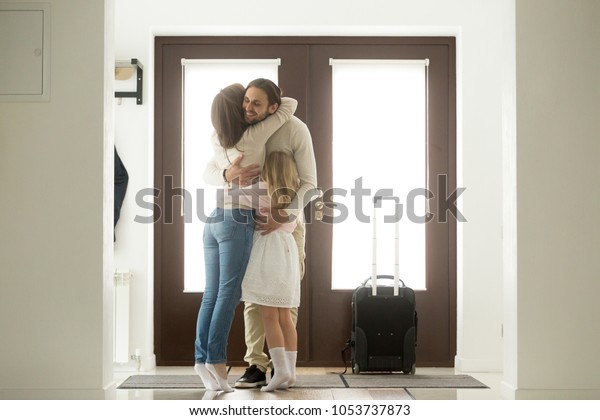 Happy dad hugging family tight arriving from long\
business trip with suitcase, smiling loving father embracing wife\
and daughter standing in house hall, welcome back home daddy and\
reunion concept