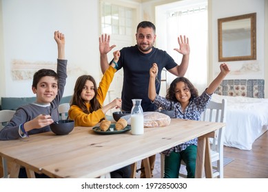 Happy dad with kids sitting in kitchen, rising their hands up. Kids enjoying their delicious breakfast meal. Breakfast, fatherhood, family concept - Shutterstock ID 1972850030