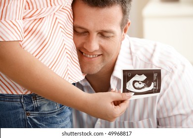 Happy Dad Listening To Pregnant Wife's Tummy, Smiling, Woman Holding Ultrasound Baby Photo.