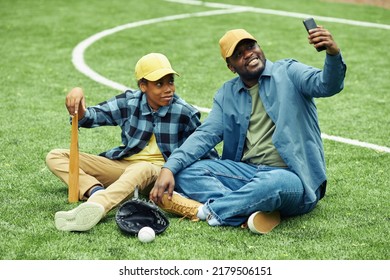 Happy Dad Making Selfie Portrait With His Son On His Mobile Phone While They Resting On Grass After Baseball Game