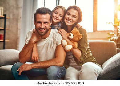 Happy Dad And Mom With Their Cute Daughter And Teddy Bear Hug And Have Fun Sitting On The Sofa In The Living Room At Home.
