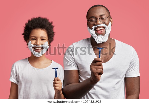 Happy dad and son with pleasant appearance, have\
shaving foam on faces, hold razors and going to shave, stand in\
frot of mirror in pink bathroom, have fun together. African kid\
imitates father