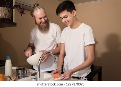 Happy Dad And Son Teenager On Kitchen. Smiling Father Cooking, Son Washing The Dishes. Concept Of Friendly Relationship Between Parents And Children Teens