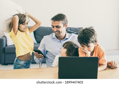 Happy Dad Using Smartphone, Sitting At Table And Kids Playing With Him. Caucasian Father Working At Home, Using Laptop And Watching Children. Fatherhood, Childhood And Digital Technology Concept