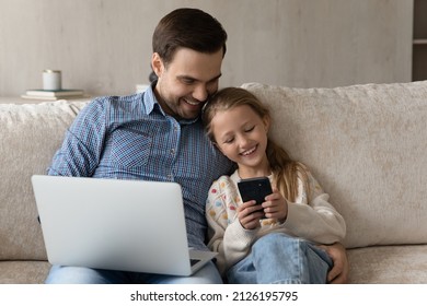 Happy daddy and gen Z daughter kid using smartphone and laptop, relaxing on sofa together, talking, laughing, watching media content. Dad teaching girl to use app on internet. Family with gadgets