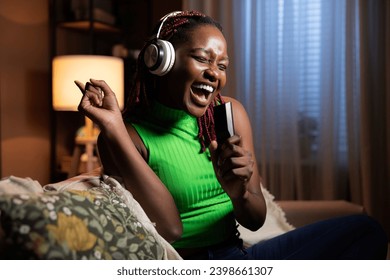 Happy dark skinned afro american woman waring modern new headphone smiling singing in remote control as microphone in casual outfit at home alone resting relaxing while sitting at home. - Powered by Shutterstock