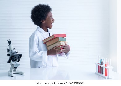 Happy Dark Skinned Boy Wear White Lab Coat Holding Lots Of Books On Arm Near Microscope In Laboratory