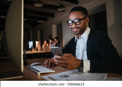 Happy dark skinned businessman working late at night in office with computer. Chatting by phone.