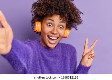 Happy Dark Skinned Female Makes Photo Of Herself Shows Peace Gesture Smiles Broadly Wears Wireless Stereo Headphones Wears Cashmere Jumper Poses Against Purple Background Has Fun Listens Music