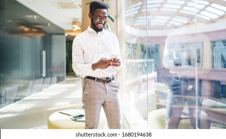Happy dark skinned man in white shirt laughing while using cellphone device for checking business news on website,cheerful male employee in classic glasses browsing wireless internet via mobile gadget