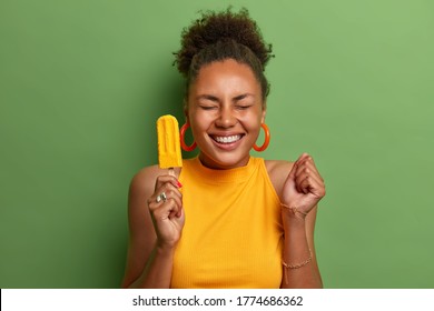 Happy dark skinned millennial girl eats yellow ice cream of mango flavor, clenches fist with anticipation of something awesome happen, has toothy smile, wears bright summer clothes, isolated on green