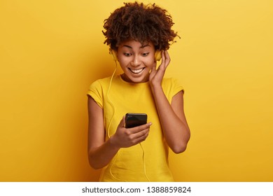 Happy dark skinned woman feels good while listens pleasant melody in headphones, focused in screen of smartphone, wears casual t shirt in one tone with background. People, technology, lifestyle
