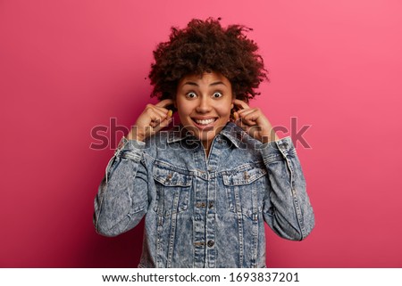 Happy dark skinned woman plugs ears, hears very loud sound, comes on very noisy party, smiles pleasantly, wears fashionable denim jacket, isolated on pink wall. People, decibel, loud music, voice