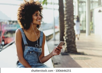 a happy dark-skinned girl enjoys music from her playlist through headphones. girl uses a mobile phone to watch a new video of her favorite artist. Female person listening online radio via cellphone - Powered by Shutterstock