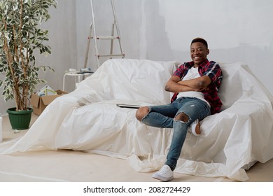 A Happy, Dark-skinned Man Sits On A Couch Covered With Plastic Sheeting In New Apartment During Renovations, The Boy Is Proud Of The Move, Smiling With Arms Crossed Over His Chest