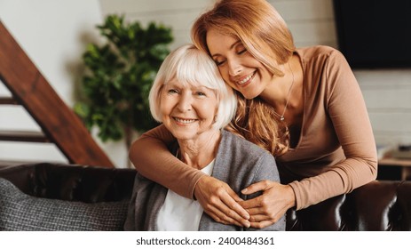 Happy daughter embracing from behind elderly mother at living room. Love and care. Family time and bonding concept. Assistance and support of elder generation - Powered by Shutterstock