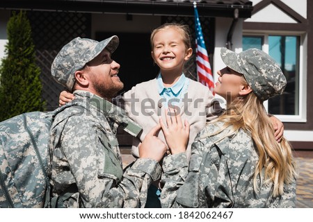 Happy daughter embracing father and mother in military uniforms near house with usa flag