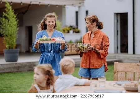 Happy daughter and mother bringing salad and burgers at multi generation garden party in summer.