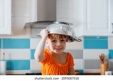 Happy Daughter Portrait Playing In Kitchen. Child Kid Girl Soiled In Flour Put Colander Bowl On Head Like Hat Have Fun And Laughs Play Game. Happy Jokes Fun While Cooking Time