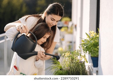 Happy daughter watering plants with her beautiful mom. Girl's in bright overall gardening on balcony - Powered by Shutterstock