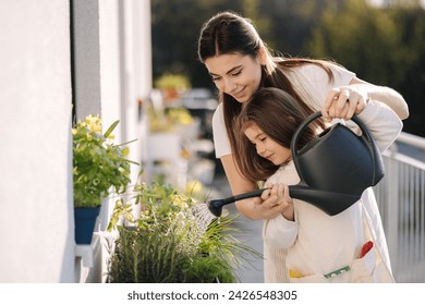 Happy daughter watering plants with her beautiful mom. Girl's in bright overall gardening on balcony - Powered by Shutterstock