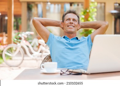 Happy day dreamer. Relaxed mature man holding hands behind head and smiling while sitting at the table outdoors with laptop on it  - Powered by Shutterstock