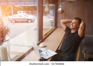 Happy day dreamer. Relaxed young man holding hands behind head and smiling while sitting at the table with laptop in modern cafe - Shutterstock ID 517345846