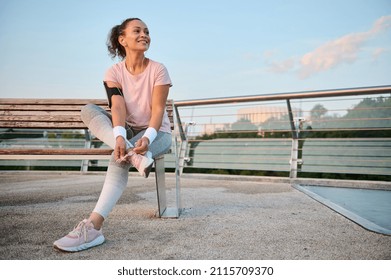 Happy delighted Caucasian female athlete sportswoman in pink t-shirt with smartphone holder and terry wristbands rests after morning jogging at dawn sitting on a wooden bench in an urban environment - Powered by Shutterstock