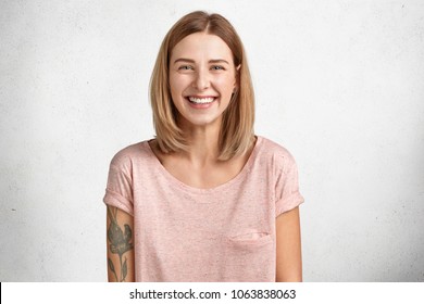 Happy delighted female with positive smile, has tattoo, smiles broadly, dressed in casual clothing, isolated over white concrete wall. Smiling adorable glad woman rejoices success and having weekends